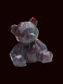A small faceted crystal bear figurine with a dark purple body and a tiny red bow on its head, sitting on a white textured tablecloth with a blurred background.