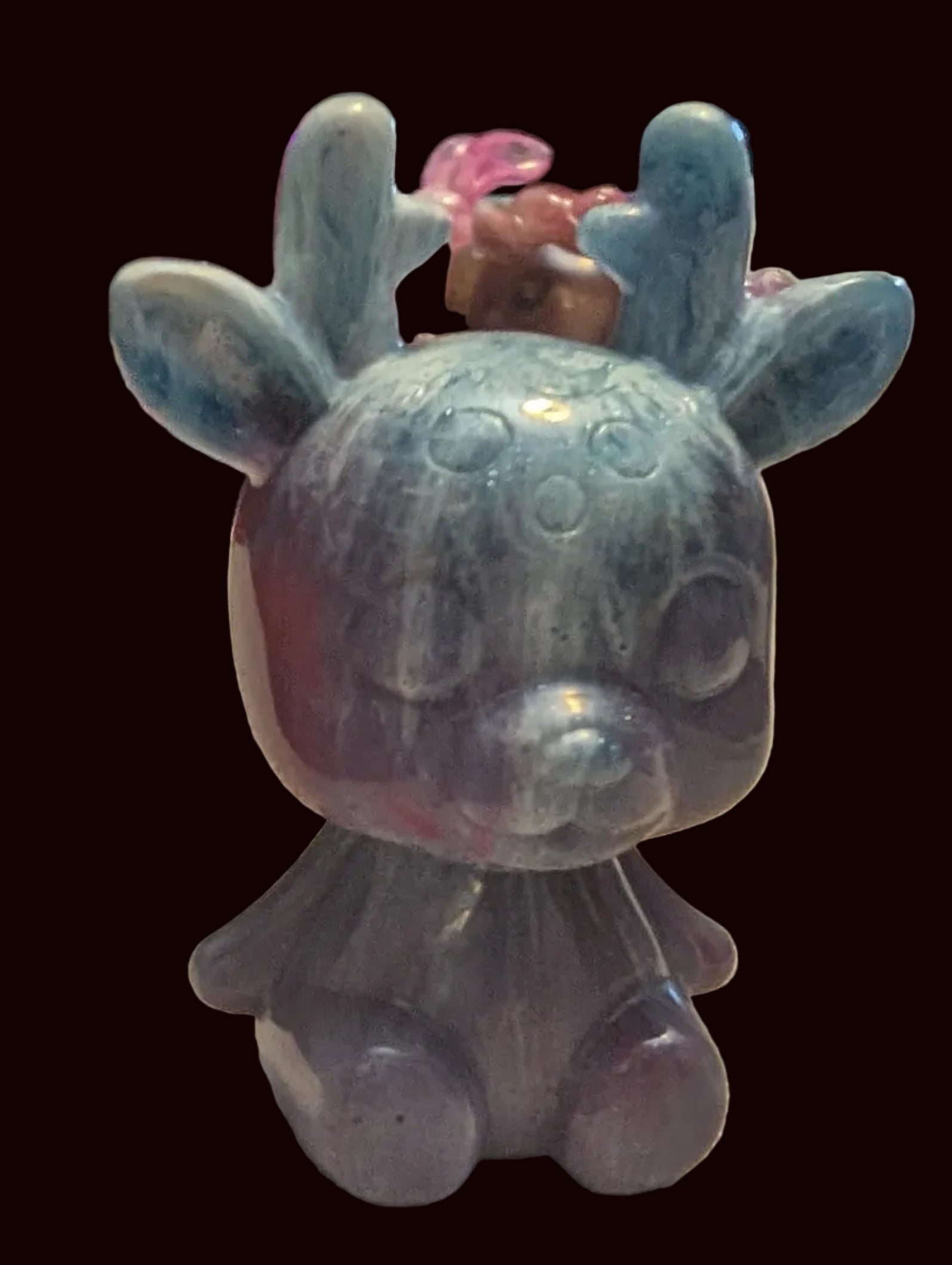 A small carved figurine of a deer or rabbit-like creature with antlers, shown sitting on a textured tablecloth in a home interior. The object has a marbled coloration with blue-green on top and purple-gray below.