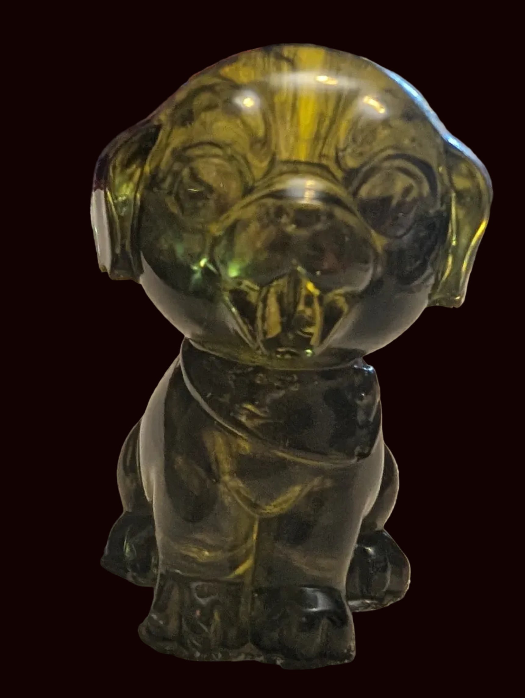 A small translucent green glass figurine of a dog sitting on a beige textured tablecloth, with a red wooden clock and other household items blurred in the background.